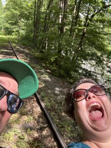 screaming out of summer (two people in the woods barely in frame as they take a selfie on a rail car ride)